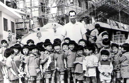 A man is surrounded by smiling children, with on-going building construction in the background.